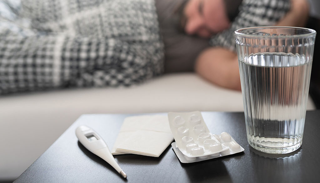 Closeup Of A Nightstand With A Glass Of Water, Medicine, & Thermometer On It & A Man Laying In Bed In The Background Prevent Flu Dehydration