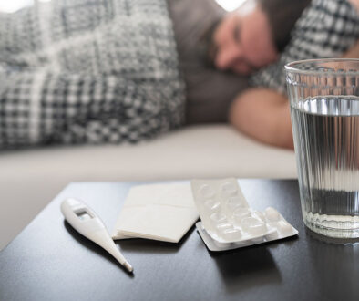 Closeup Of A Nightstand With A Glass Of Water, Medicine, & Thermometer On It & A Man Laying In Bed In The Background Prevent Flu Dehydration